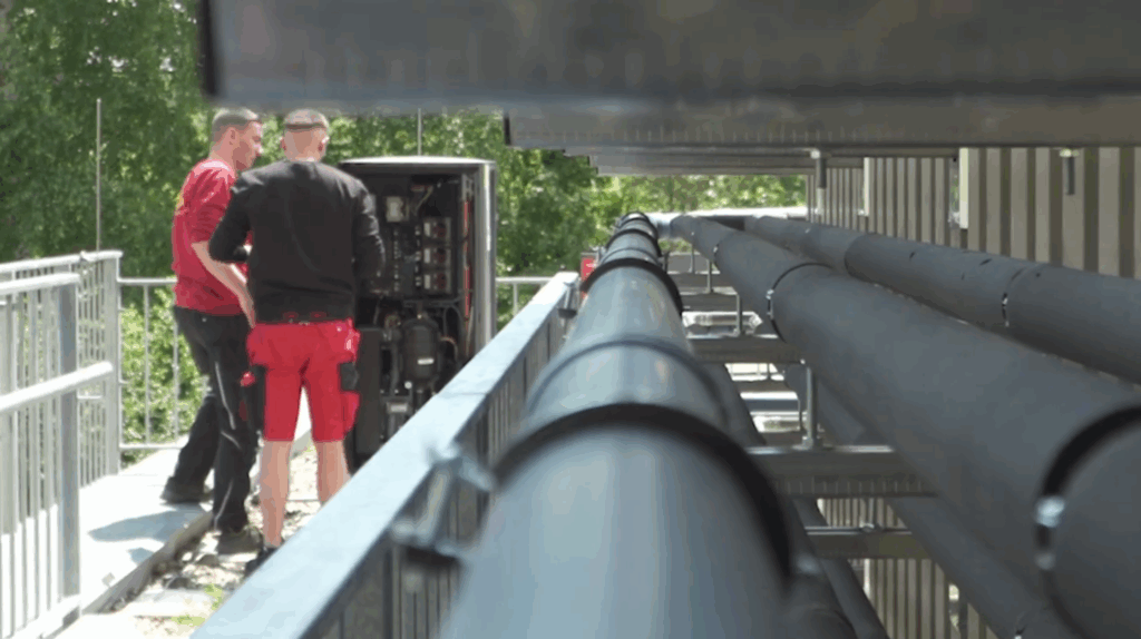 Zwei Auszubildende inspizieren und messen Komponenten an einer geöffneten Außenwärmepumpe.