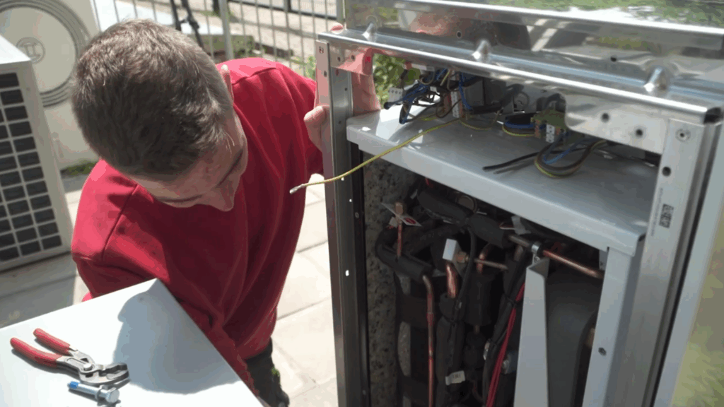 Nahaufnahme eines Auszubildenden, der an einer geöffneten Wärmepumpen-Außeneinheit Mess- und Wartungsarbeiten durchführt.